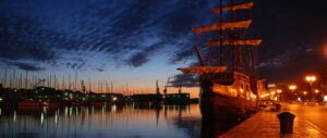 Zachód słońca nad promenadą w Trogirze w Chorwacji. Widoczna replika dawnego żaglowca i odbicia masztów jachtów stojących w marinie