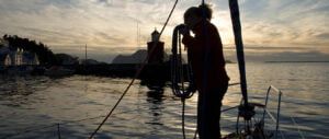 Żeglarka zwija cumę podczas rejsu morskiego po Norwegii. W tle główki port Alesund o zachodzie słońca